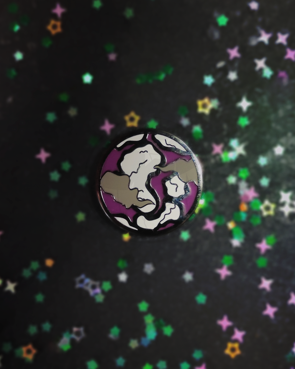 Photo of a hard enamel pin on a dark, blurry background. The pin is a stylized planet colored with the colors of the asexual flag.