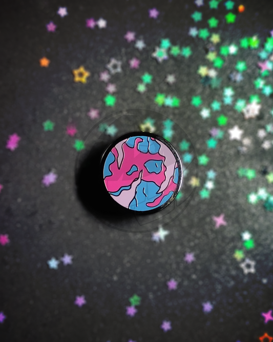 Hard enamel pin of a stylized planet on a dark, blurry background. The pin has the colors of the bisexual flag in it.