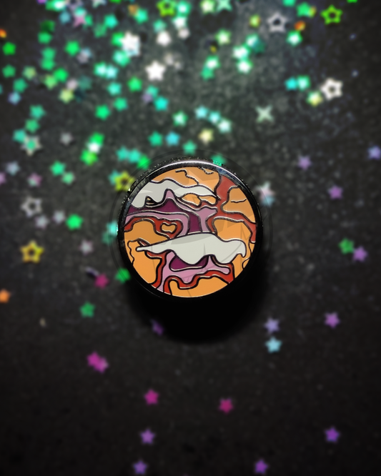 Photo of a hard enamel pin on a dark, blurry background. The pin is a stylized planet colored with the colors of the lesbian flag.