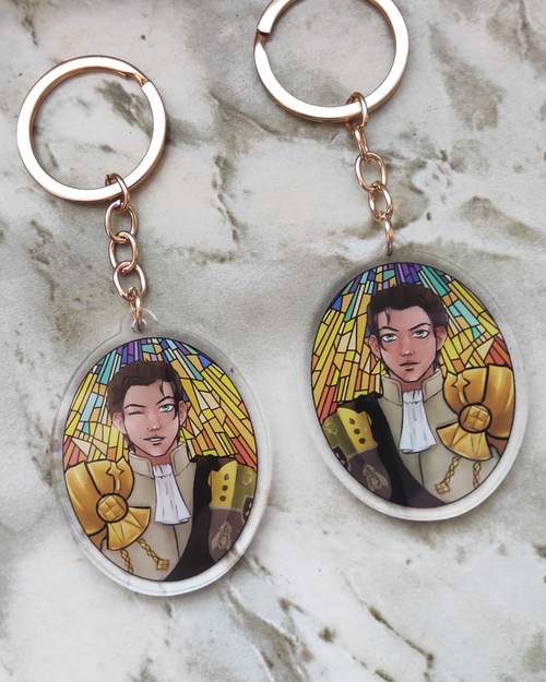Two keychains of Claude of Fire emblem on a stone background. Each side shows a different expression.